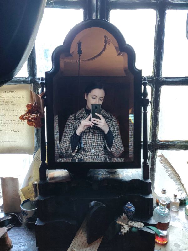 A mirror selfie of a dark haired woman in a checkered coat, standing in an old fashioned room.