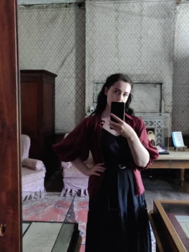 A dark haired woman in a room with peeling wallpaper, in a long black dress.
