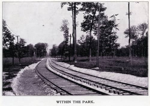 Train tracks stretching off into distant parkland. Caption says Within The Park. Old postcard.